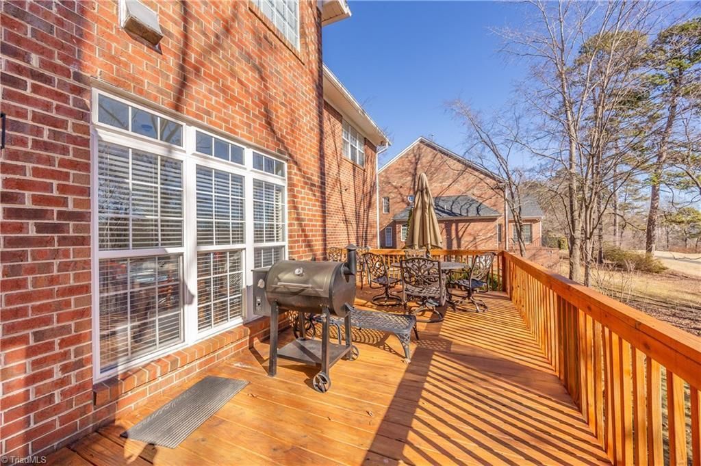 Sunlit wooden deck beside a brick house, with black grill, patio table, and orange railing.