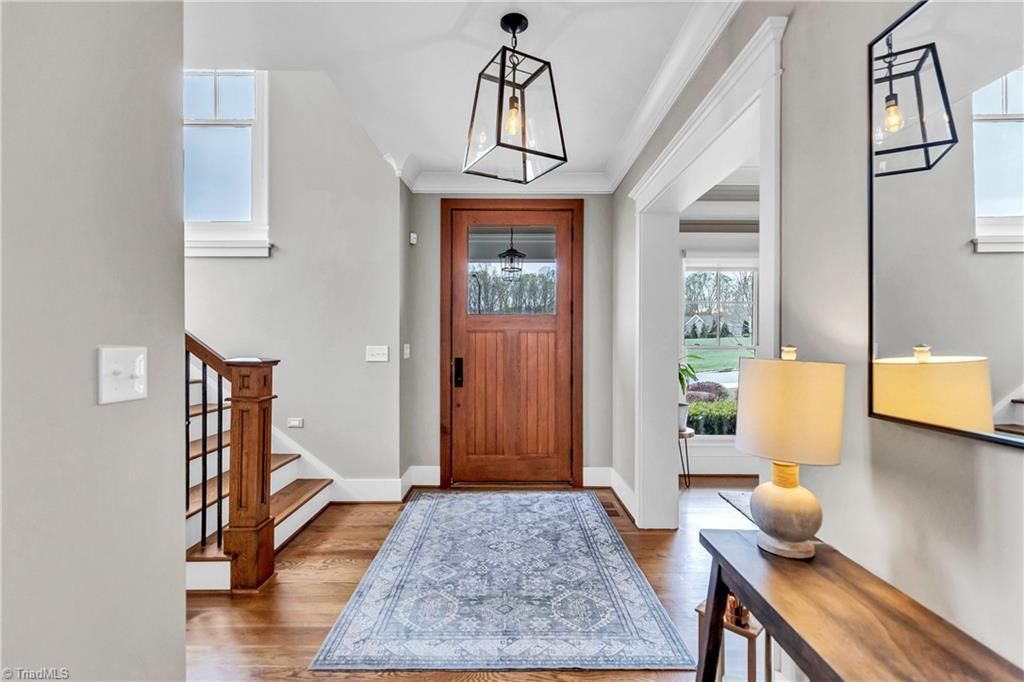 Bright entryway with wooden front door, staircase, console table, lamp, and patterned runner rug.