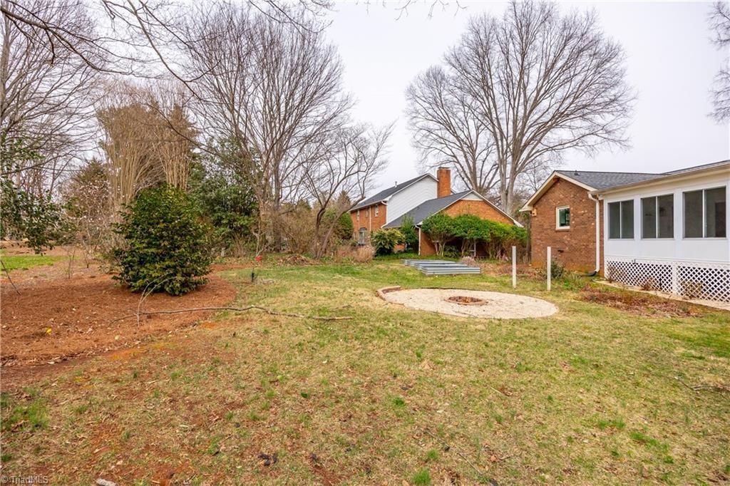 Suburban backyard with grass, trees, a small patio area, and brick houses under an overcast sky