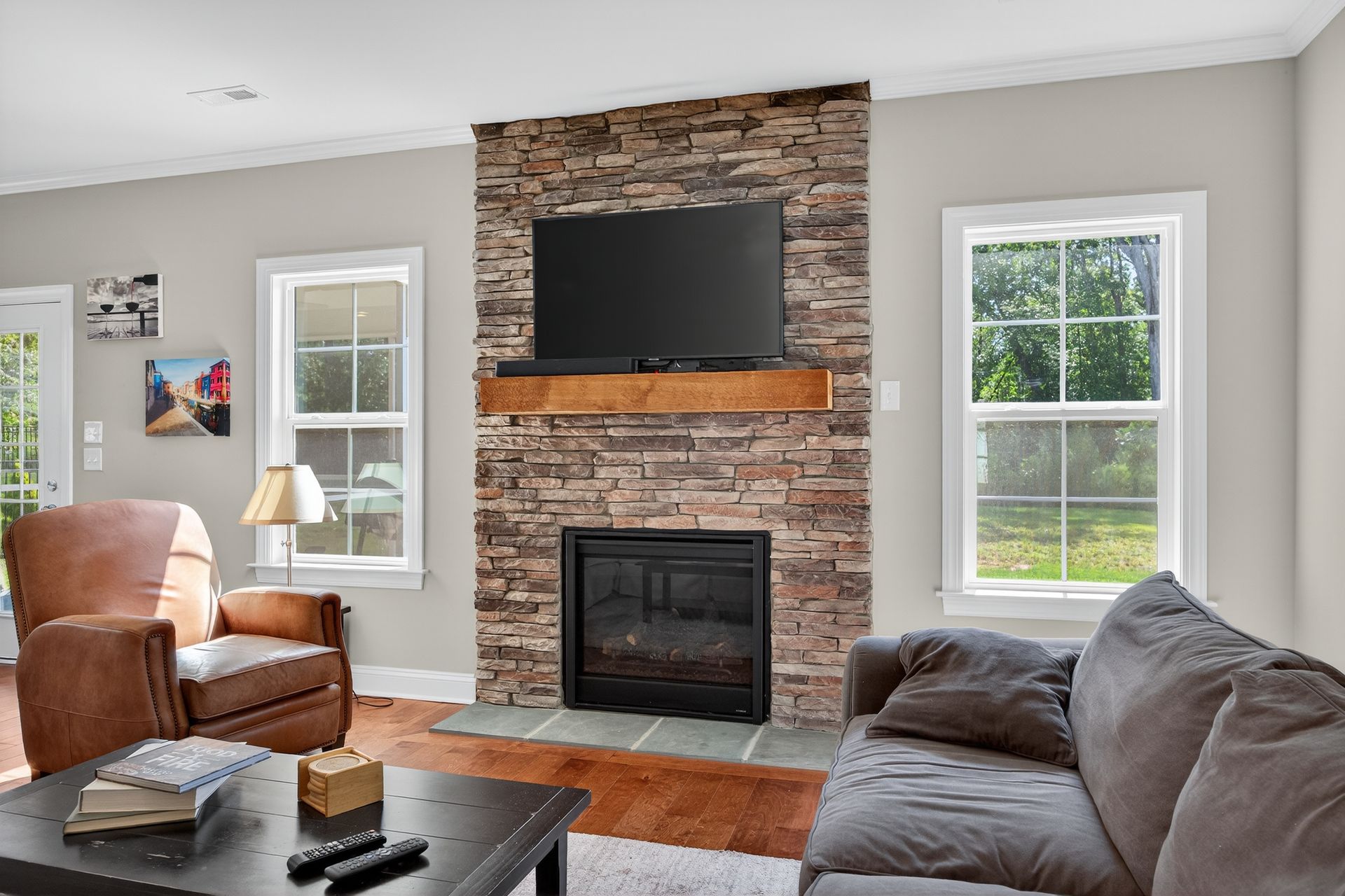 Living room with stone fireplace, mounted TV, leather chair, gray sofa, and two windows