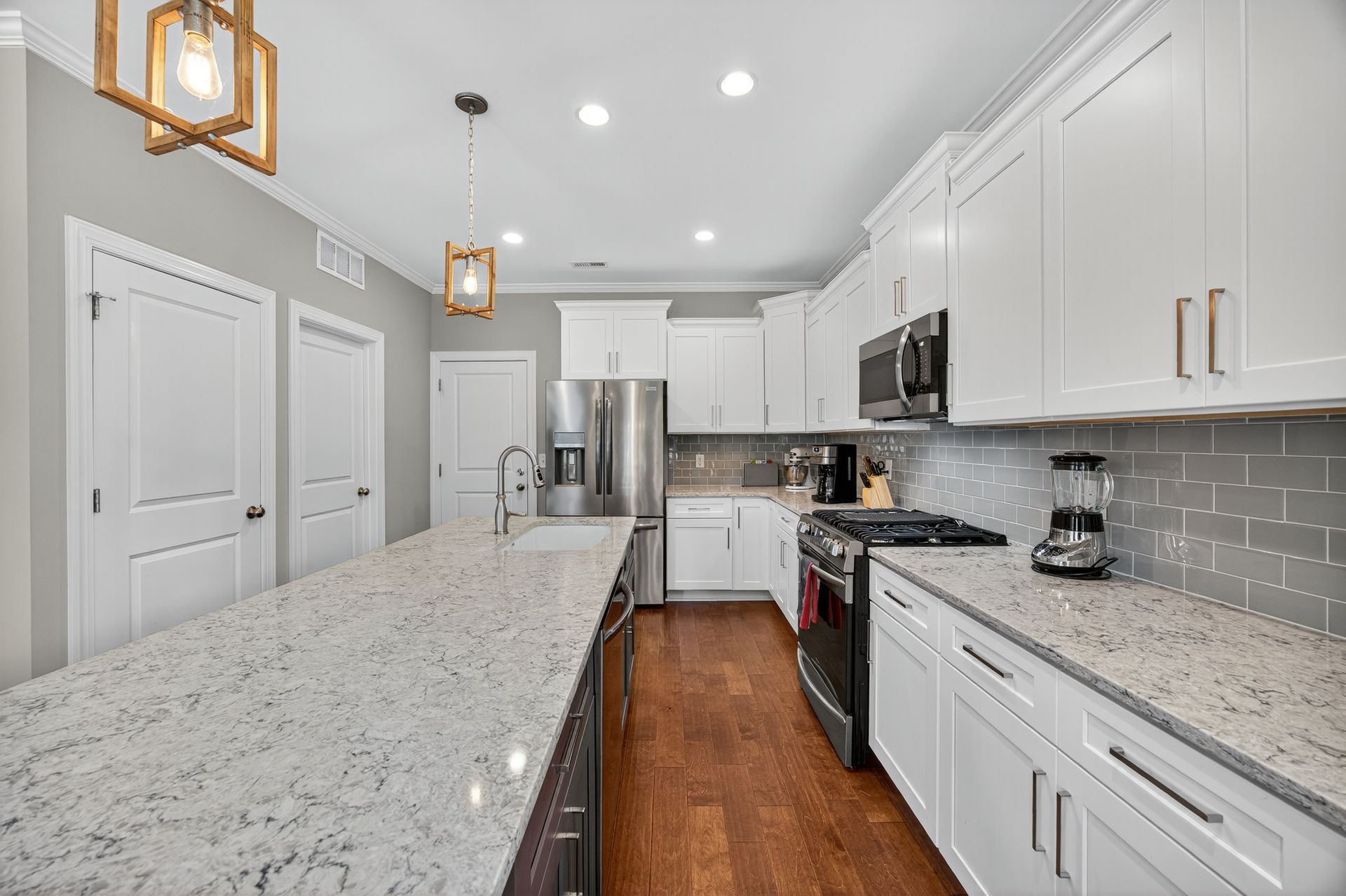 Modern white kitchen with granite island, stainless appliances, wood floor, and pendant lights.