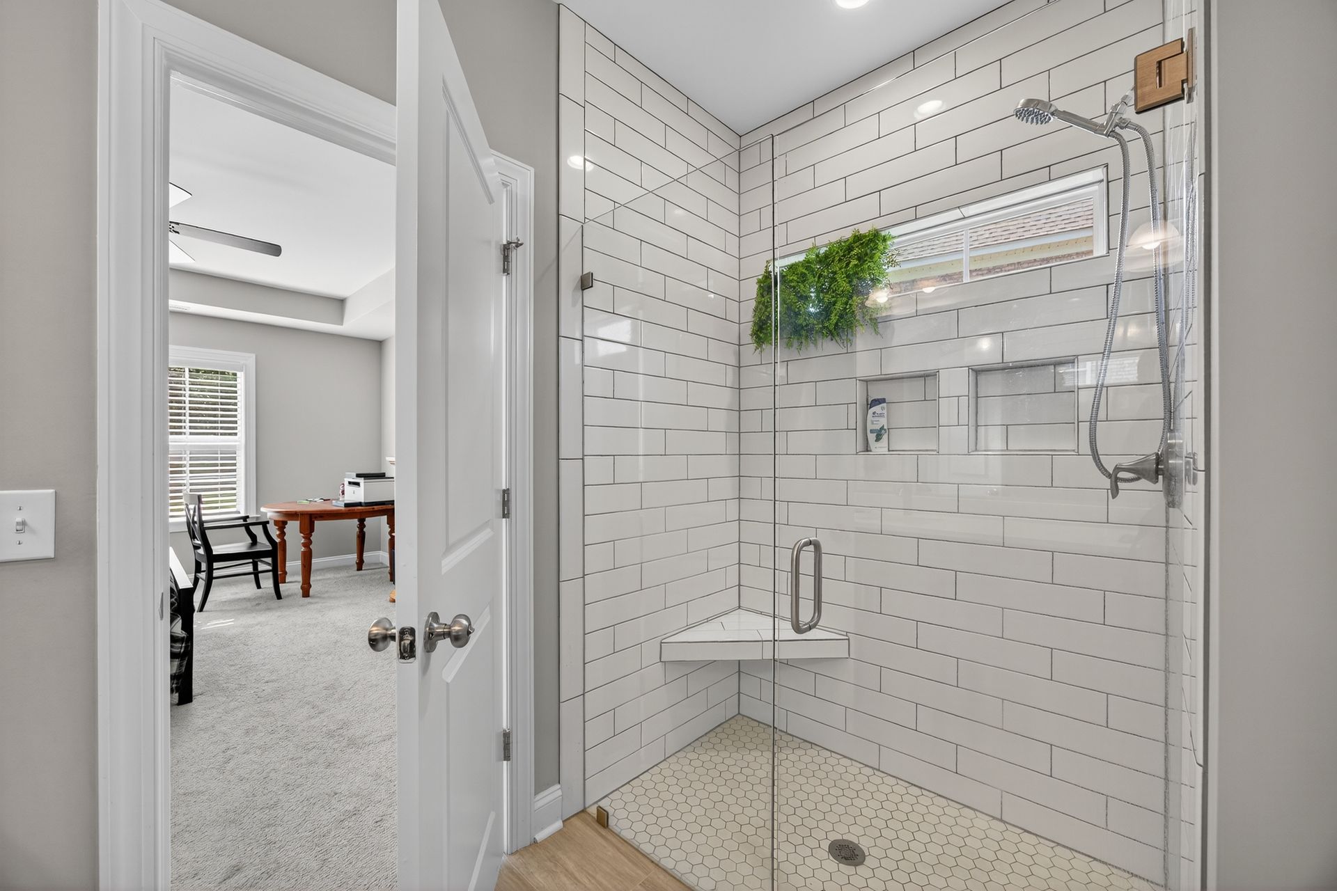 Bright walk-in shower with white subway tile, wood-look floor, glass door, and a wall shelf with greenery.