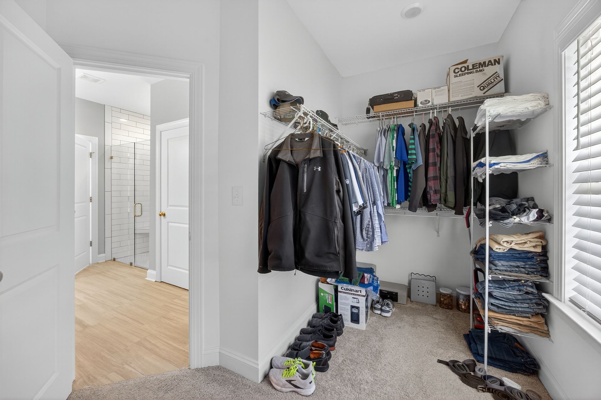 Walk-in closet with clothes on racks and shelves, beside a carpeted hallway and bathroom doorway.