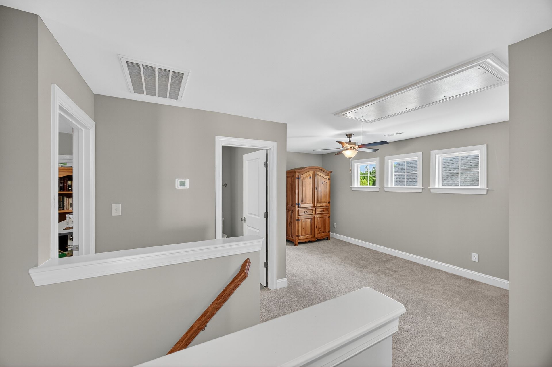 Bright carpeted living room with gray walls, stair railing, windows, and a wooden cabinet near the dining area