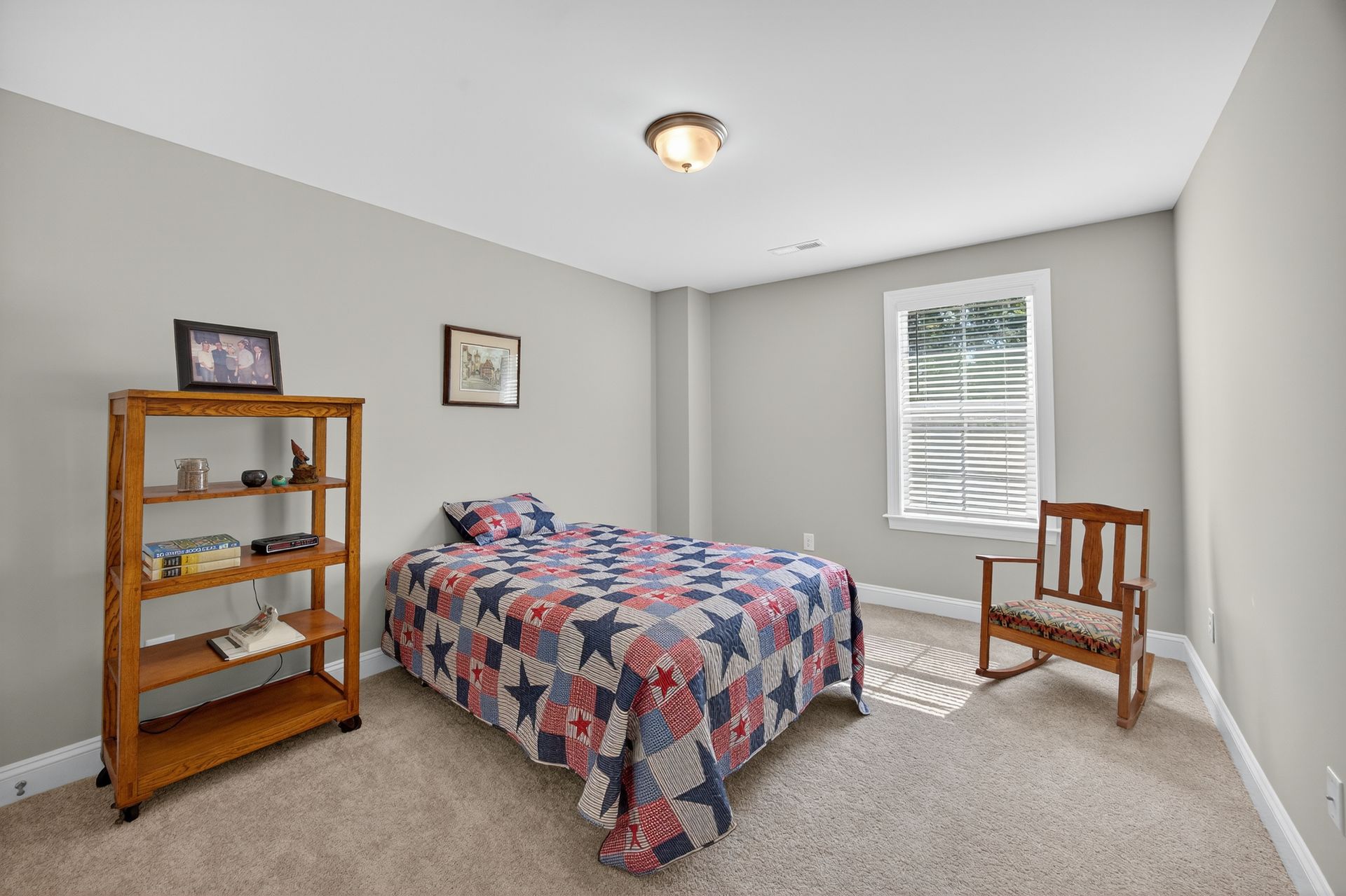 Small bedroom with floral bedspread, wooden shelf, chair, and a window with blinds