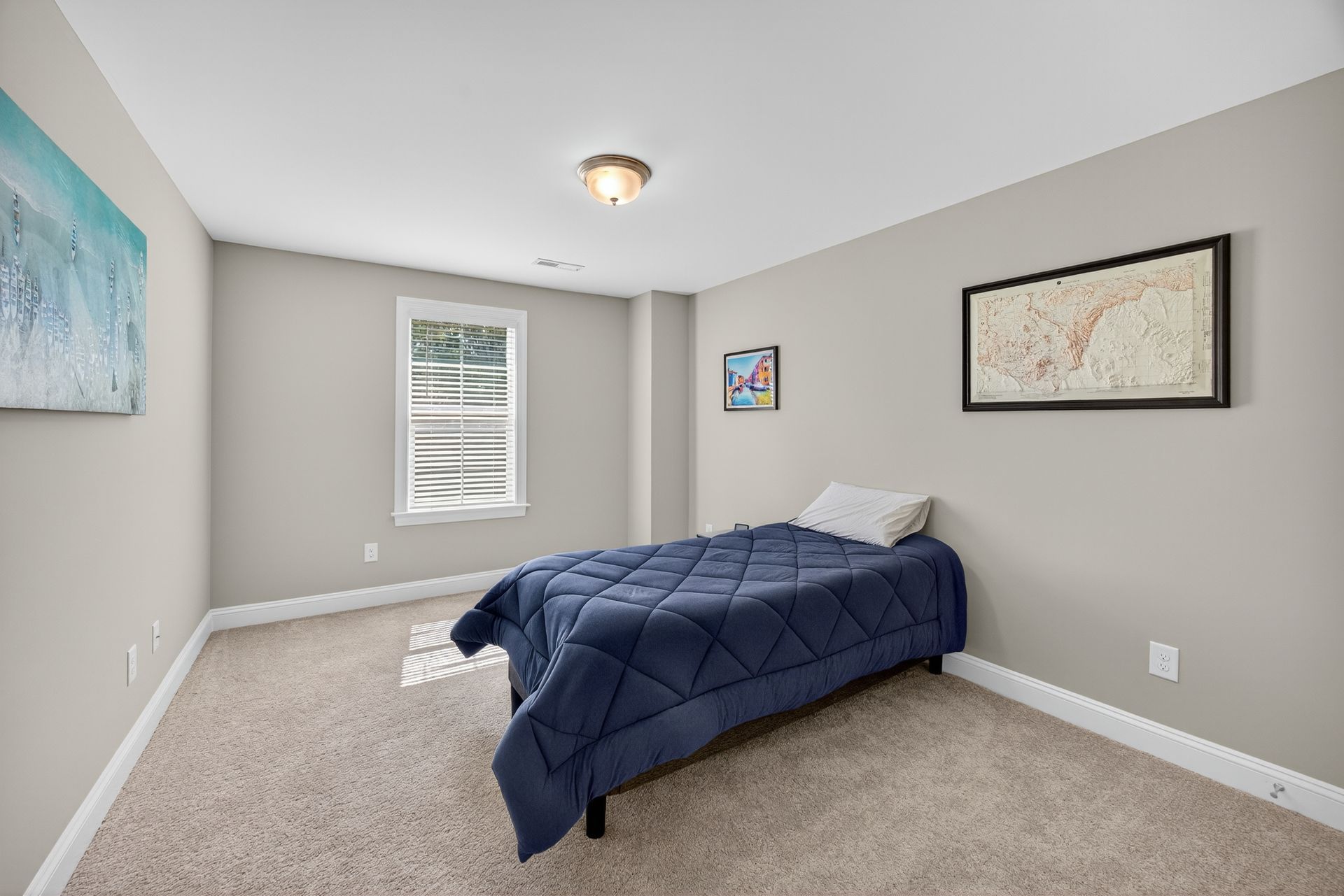 Small bedroom with a blue bedspread, beige carpet, gray walls, and wall art near a window.