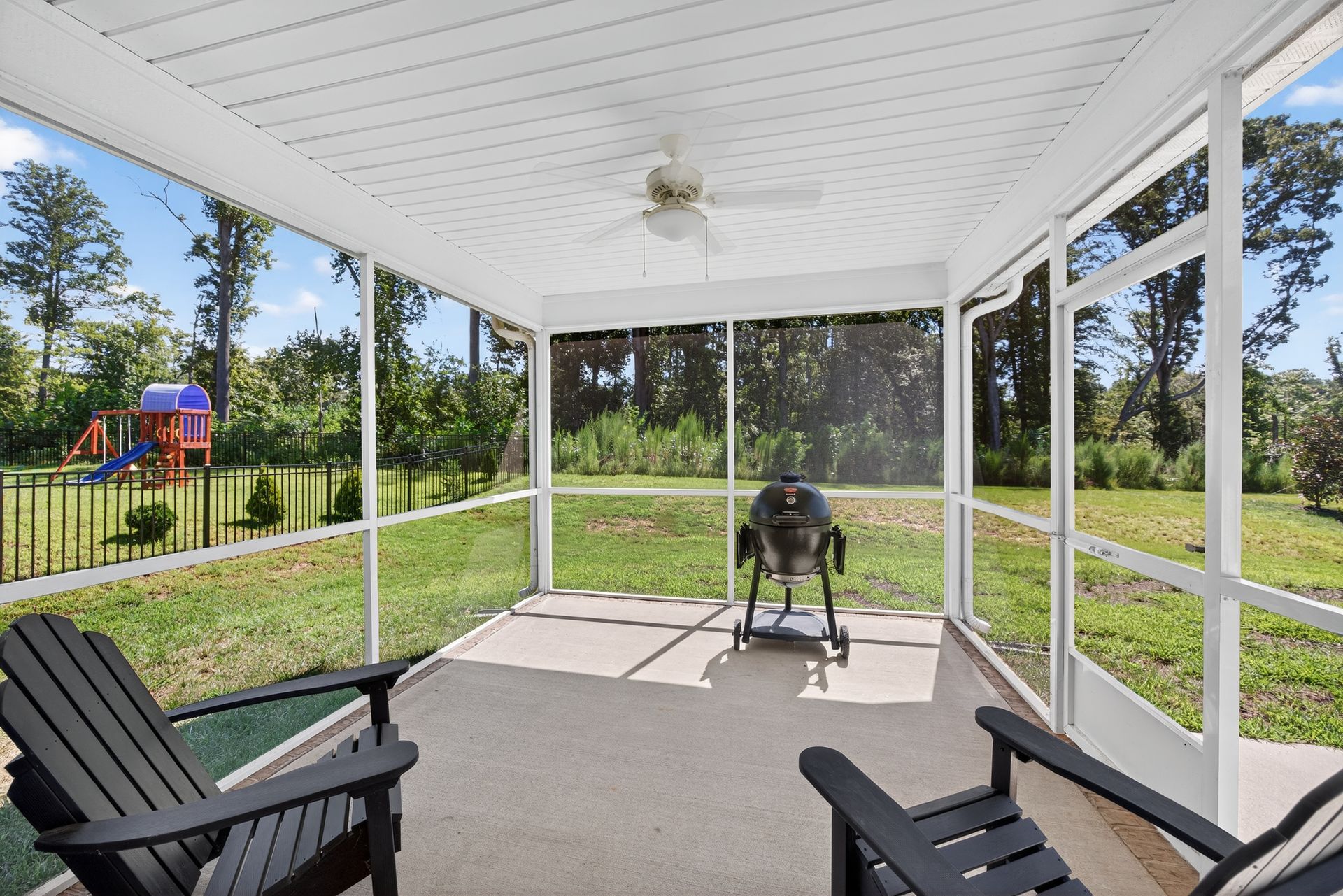 Covered screened porch with black chairs and a grill overlooking a sunny backyard and playground