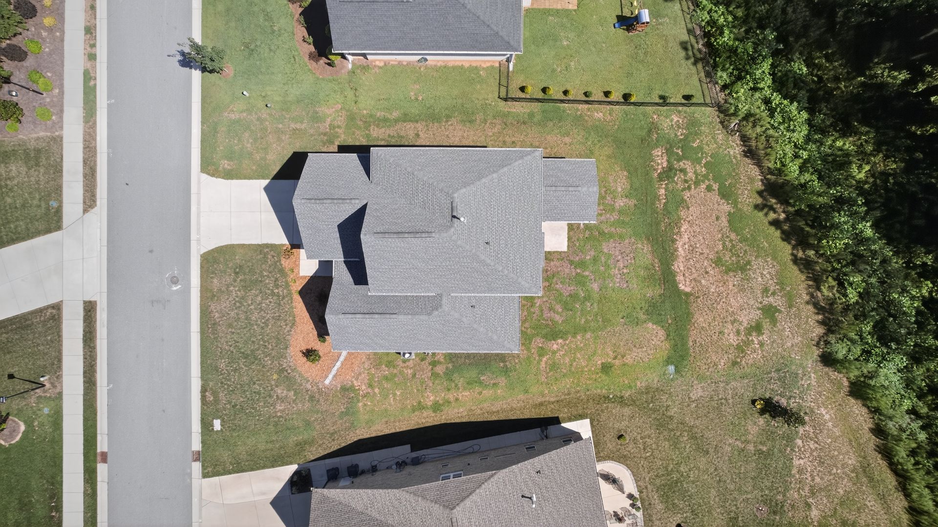 Aerial view of a house with gray roof beside a road, surrounded by grass and neighboring homes.
