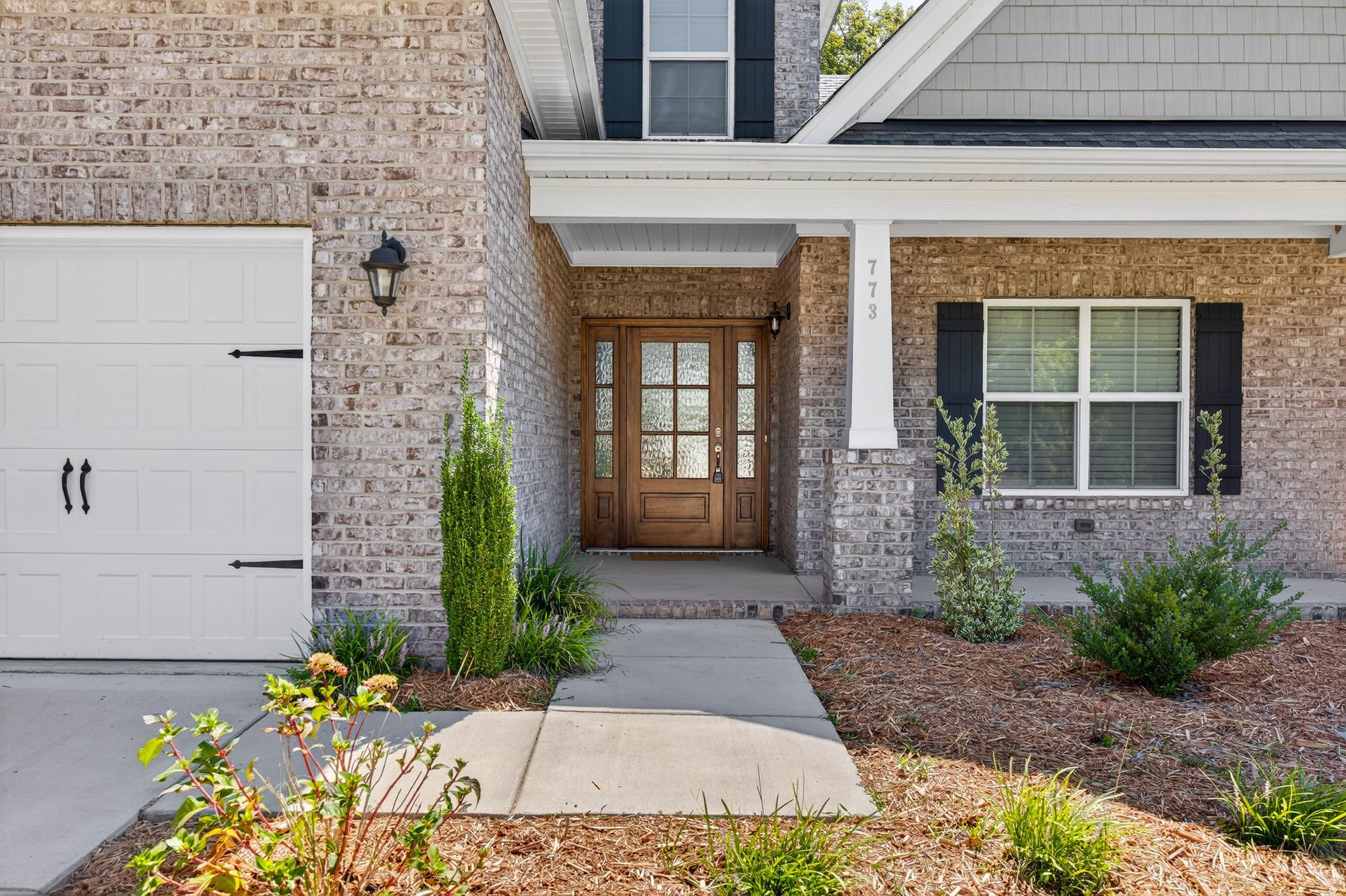 Front entrance of a brick townhouse with a white garage door, wooden front door, and landscaped walkway.