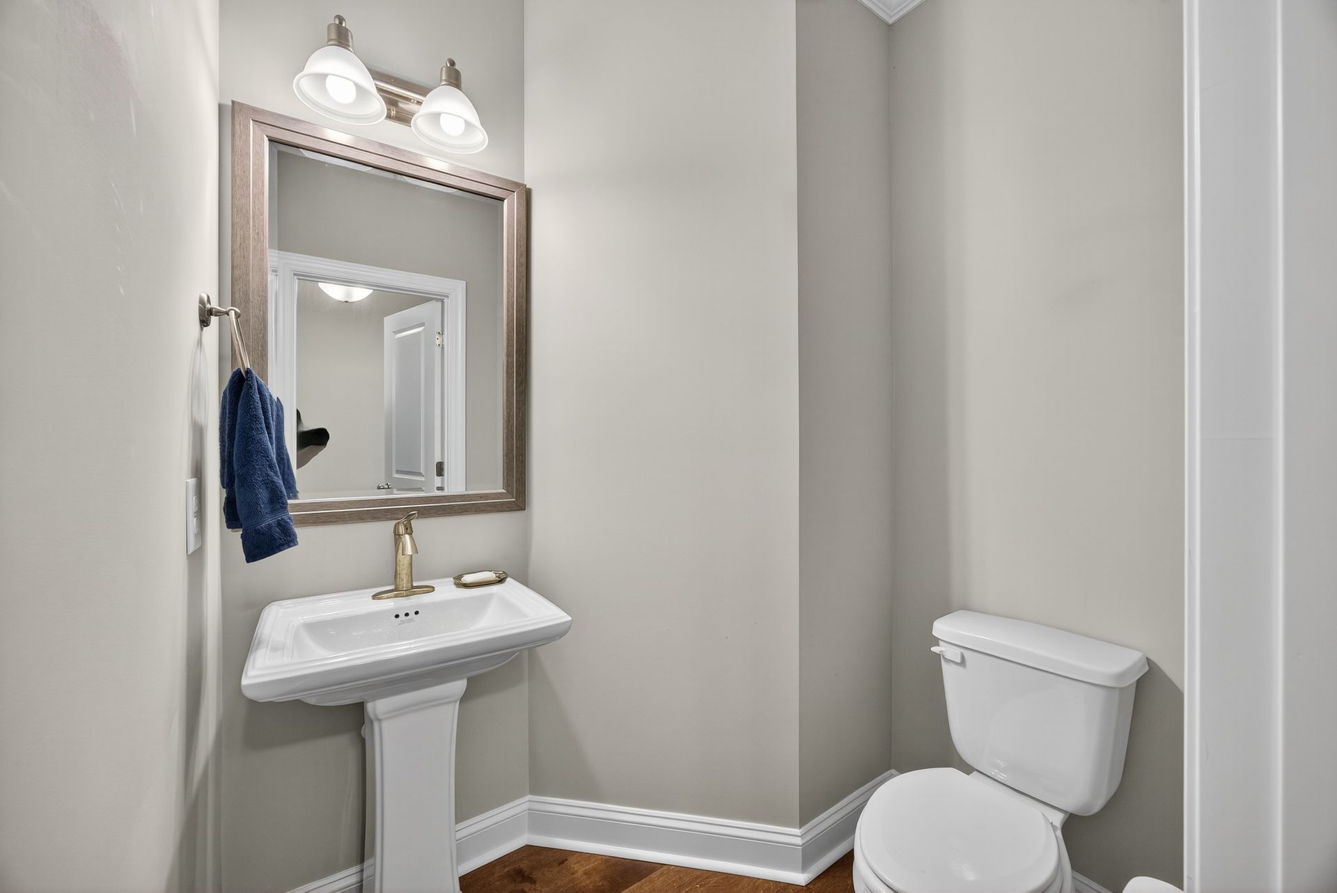 Small bathroom with pedestal sink, mirror, towel, toilet, and light wood floor.