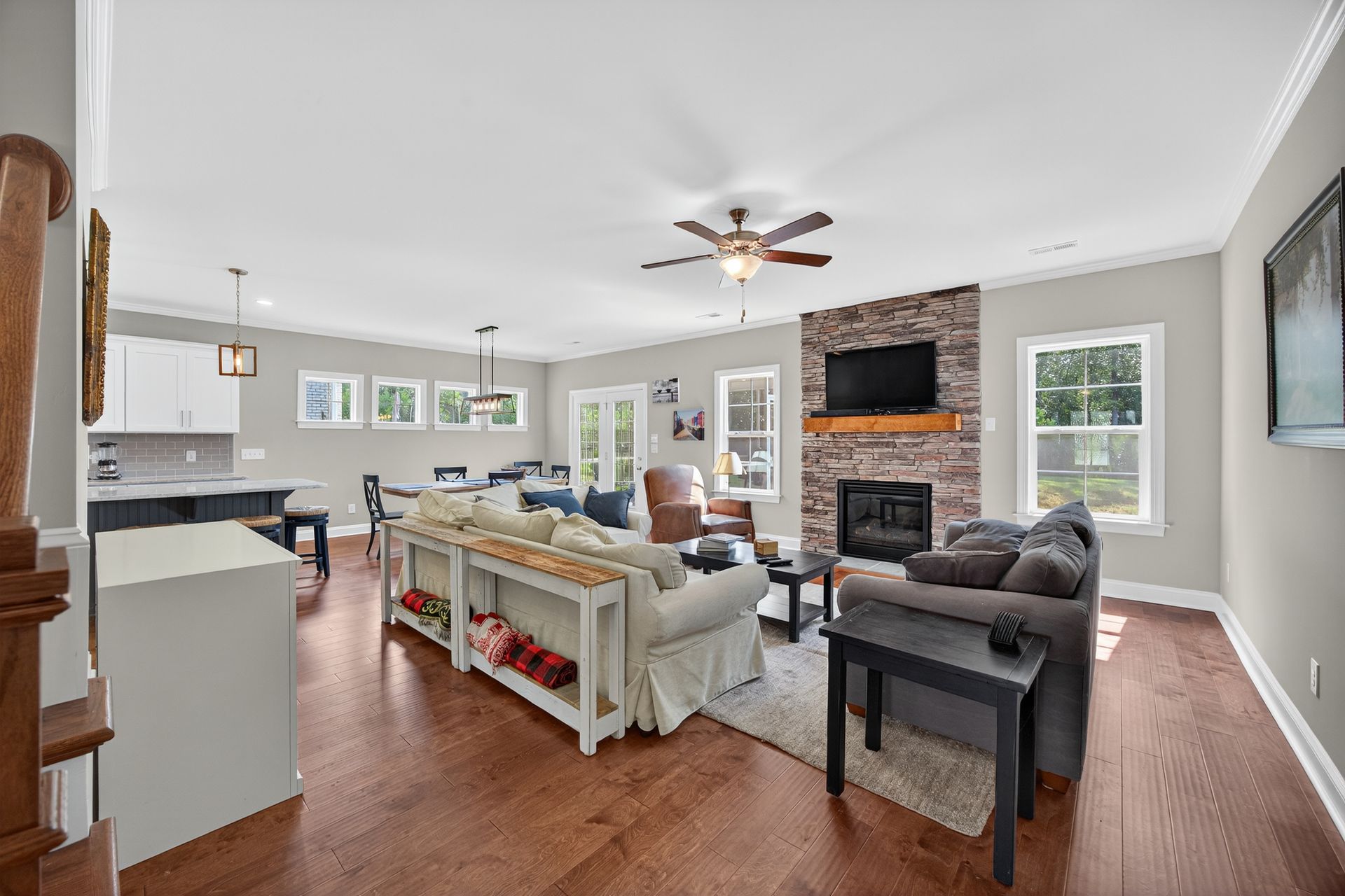 Bright open-concept living room with gray sofas, brick fireplace, ceiling fan, and kitchen bar stools.