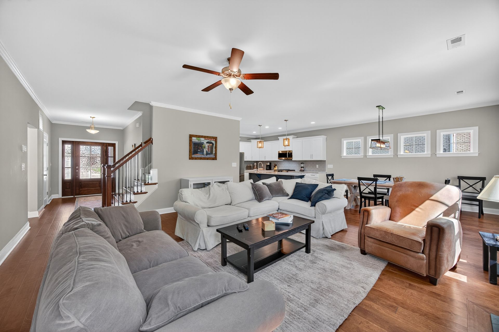 Bright open living room with gray sectional, tan chair, ceiling fan, staircase, and dining area in back