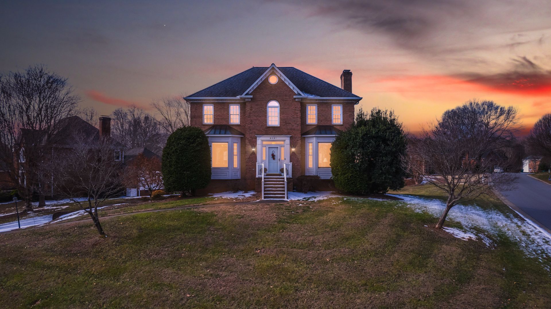 Brick house at sunset with lit porch lights, snow patches, and bare trees in the yard