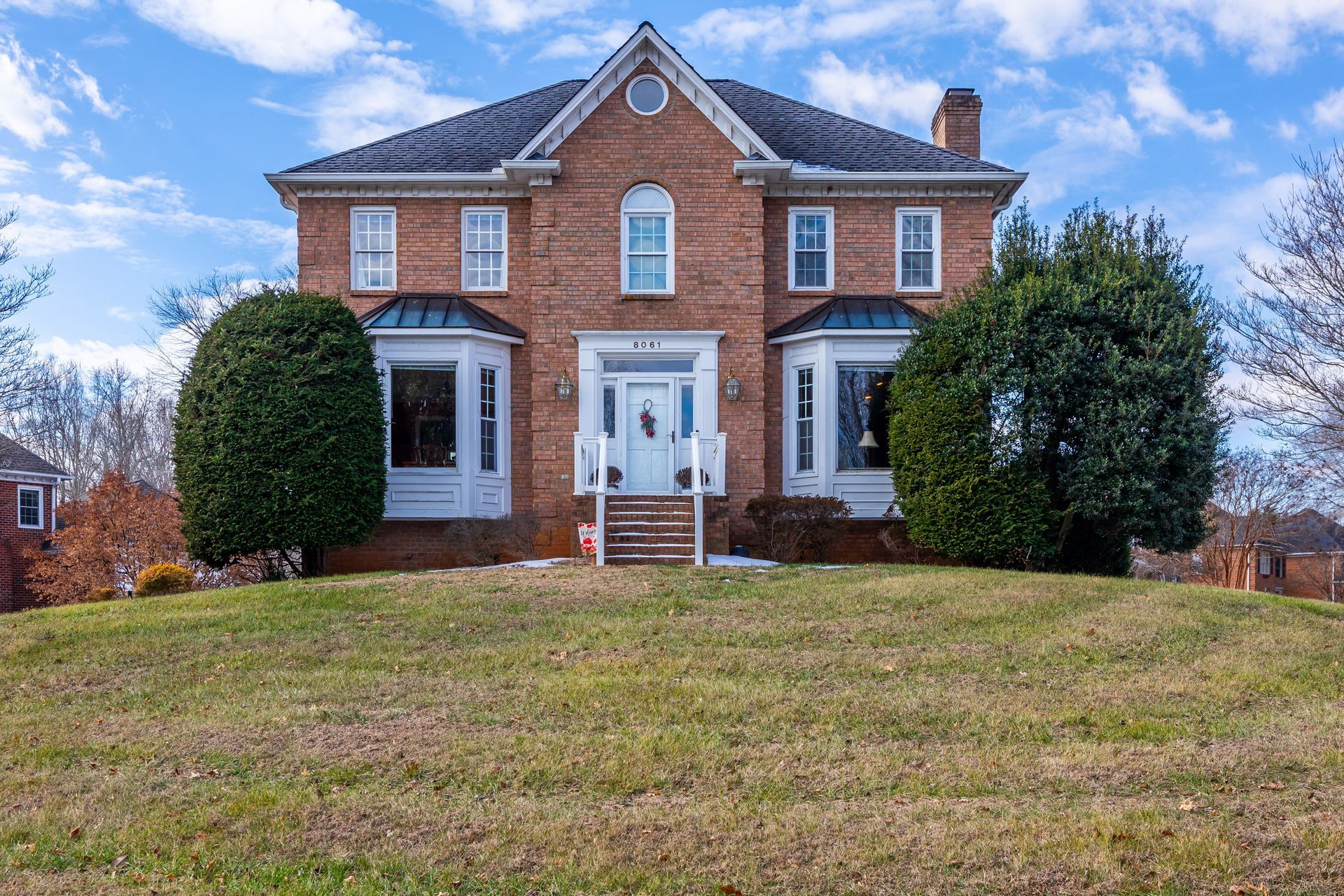 Brick house with white trim and two large shrubs, set on a grassy front lawn under a blue sky.
