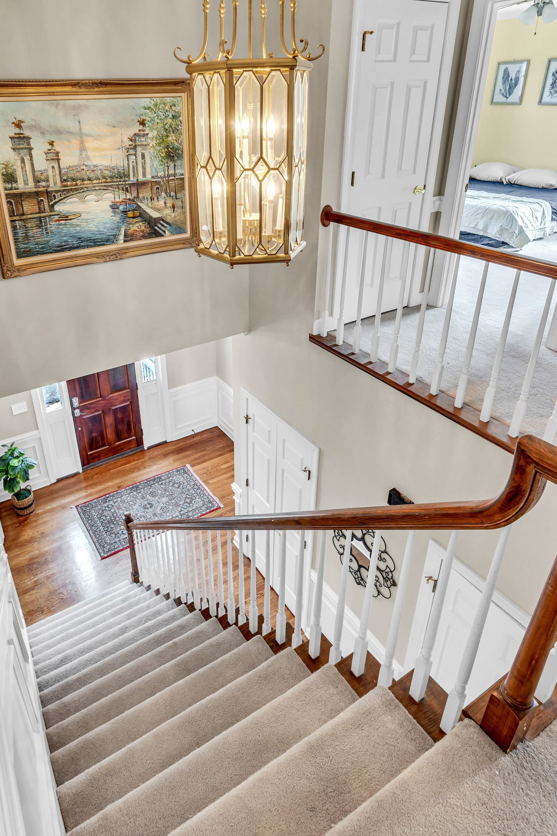 Staircase with beige carpet, white spindles, wood handrail, and a chandelier in a bright foyer