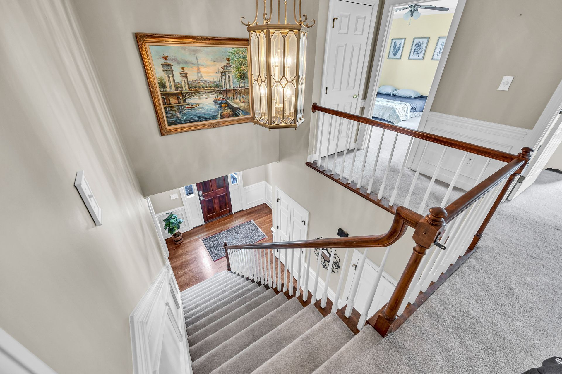 Staircase landing with white carpet, wood handrail, chandelier, and framed painting in a bright home foyer