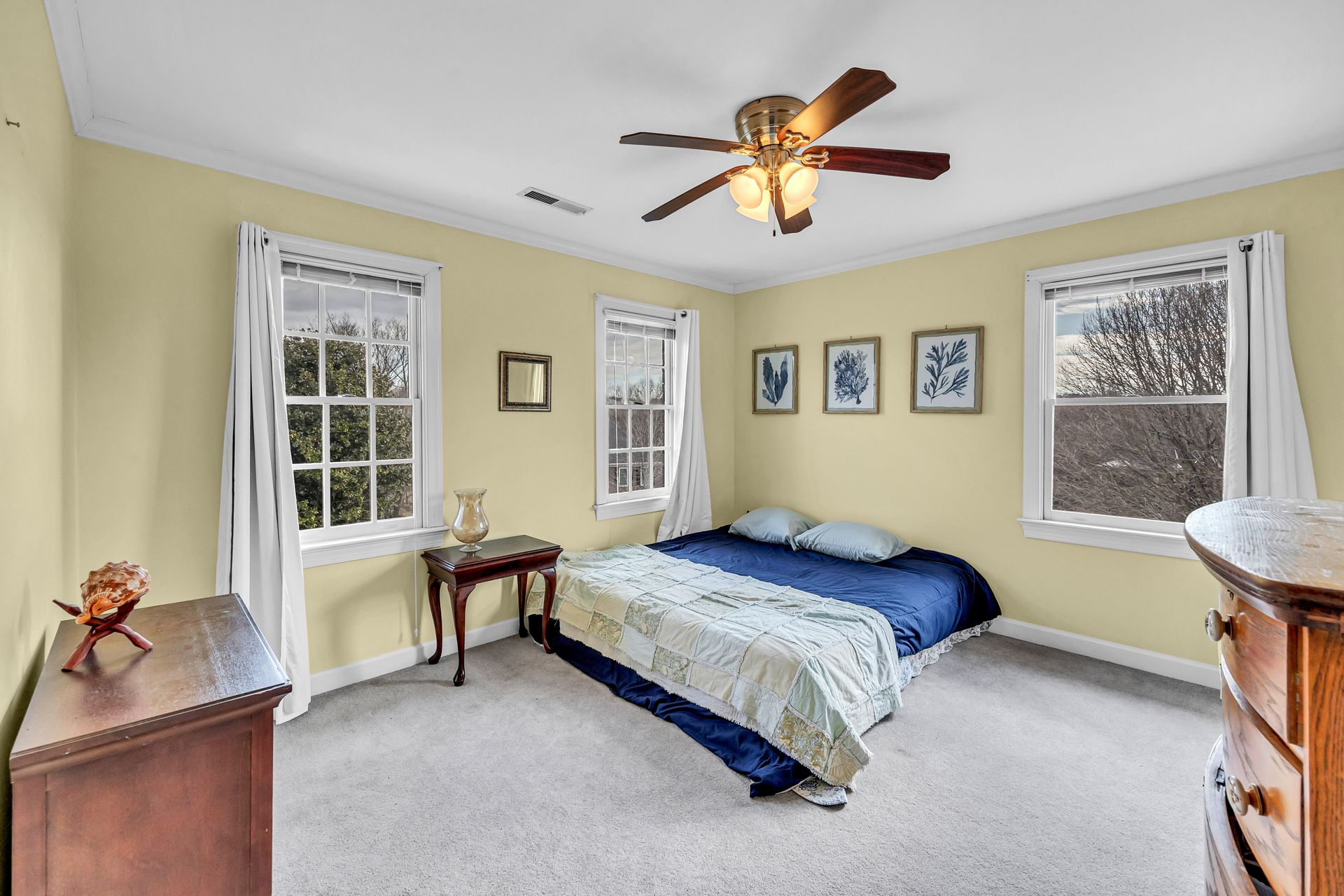 Bright bedroom with blue bedding, two windows, and a ceiling fan on a light yellow wall.