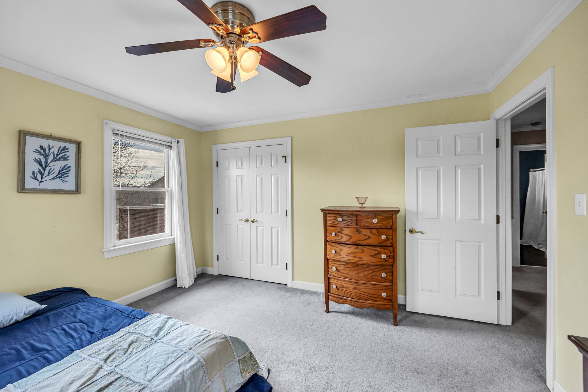 Bedroom with blue bedspread, white doors, wooden dresser, and ceiling fan on a pale yellow wall
