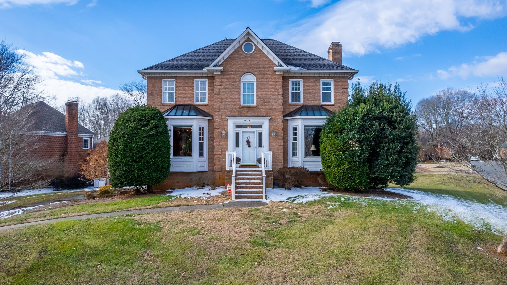 Brick two-story house with white-trim windows, front steps, and large shrubs in a winter yard