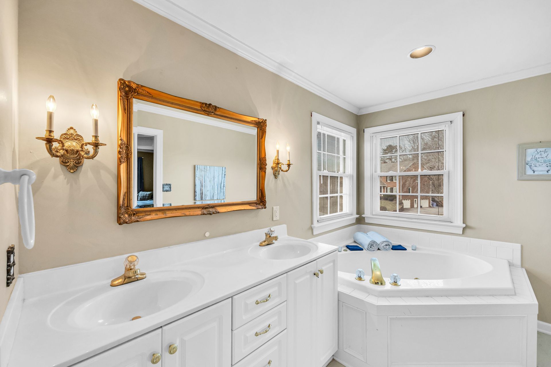 Elegant beige bathroom with double vanity, gold accents, and a corner soaking tub by a window