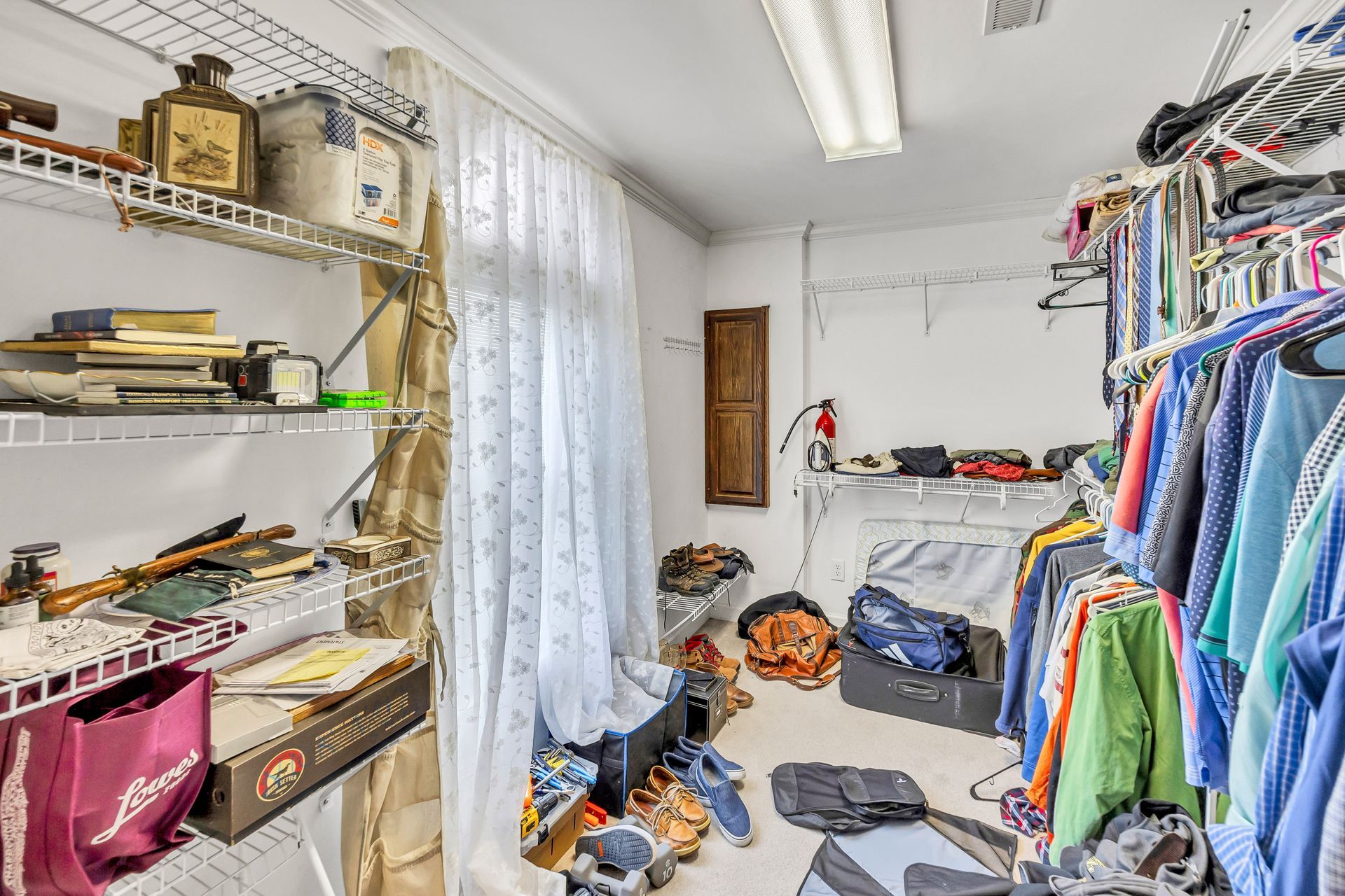 Cluttered craft room with shelves of supplies, scattered clothes, and a window with sheer curtains