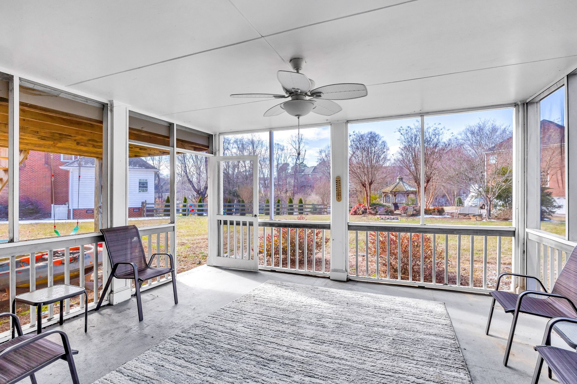 Screened porch with chairs, rug, and ceiling fan overlooking a yard and trees