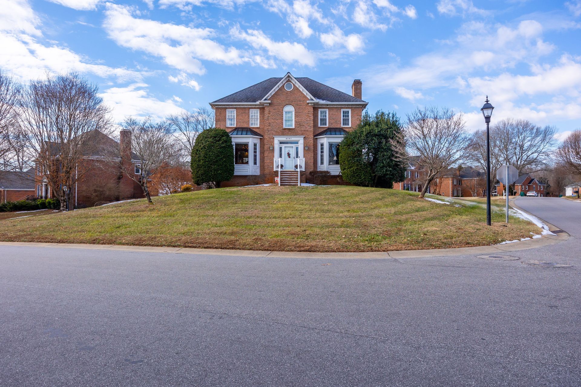 Brick house on a grassy hill beside a road, with bare trees and a cloudy blue sky.