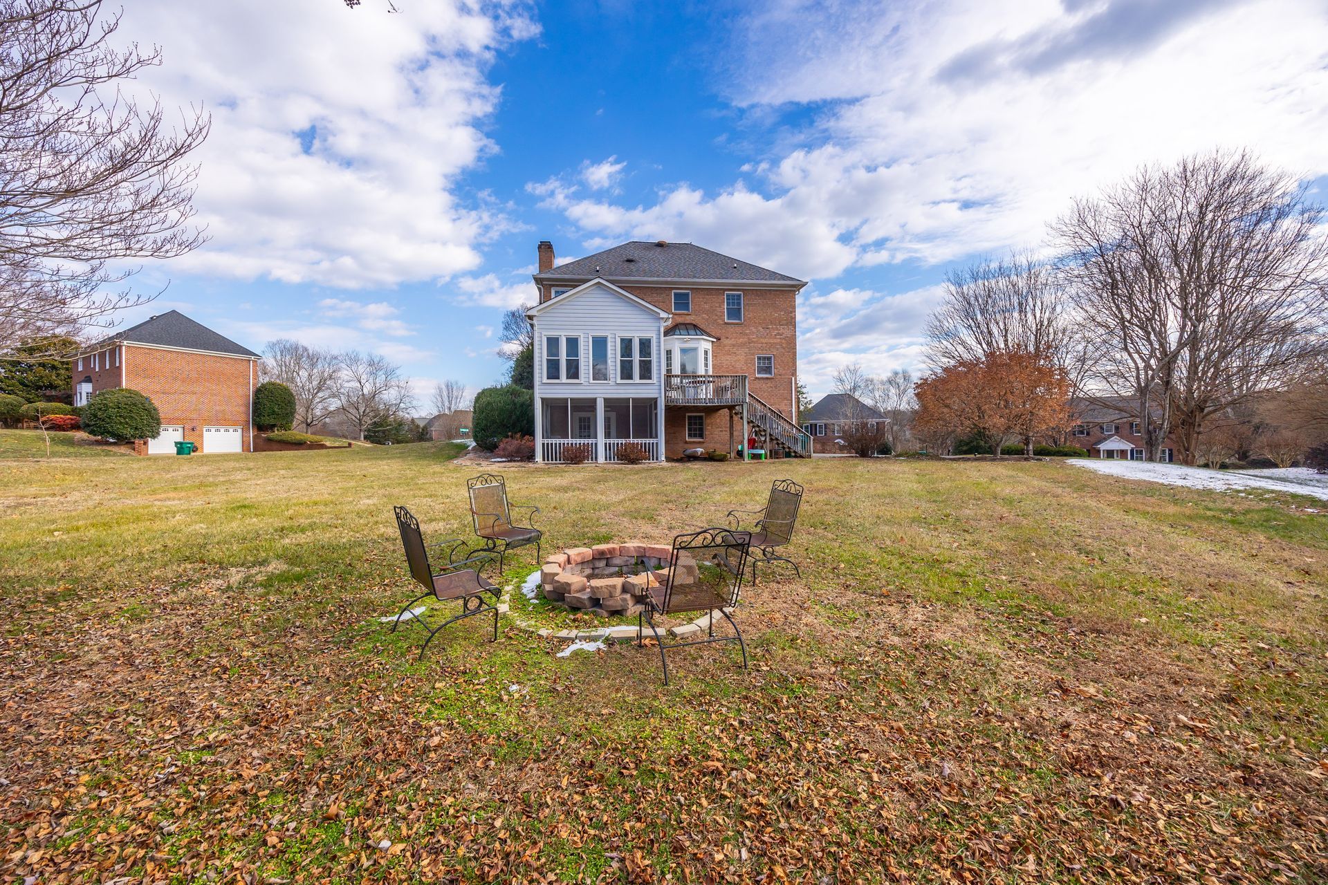 Brick house with a large backyard, patio chairs, and a fire pit under a cloudy sky.