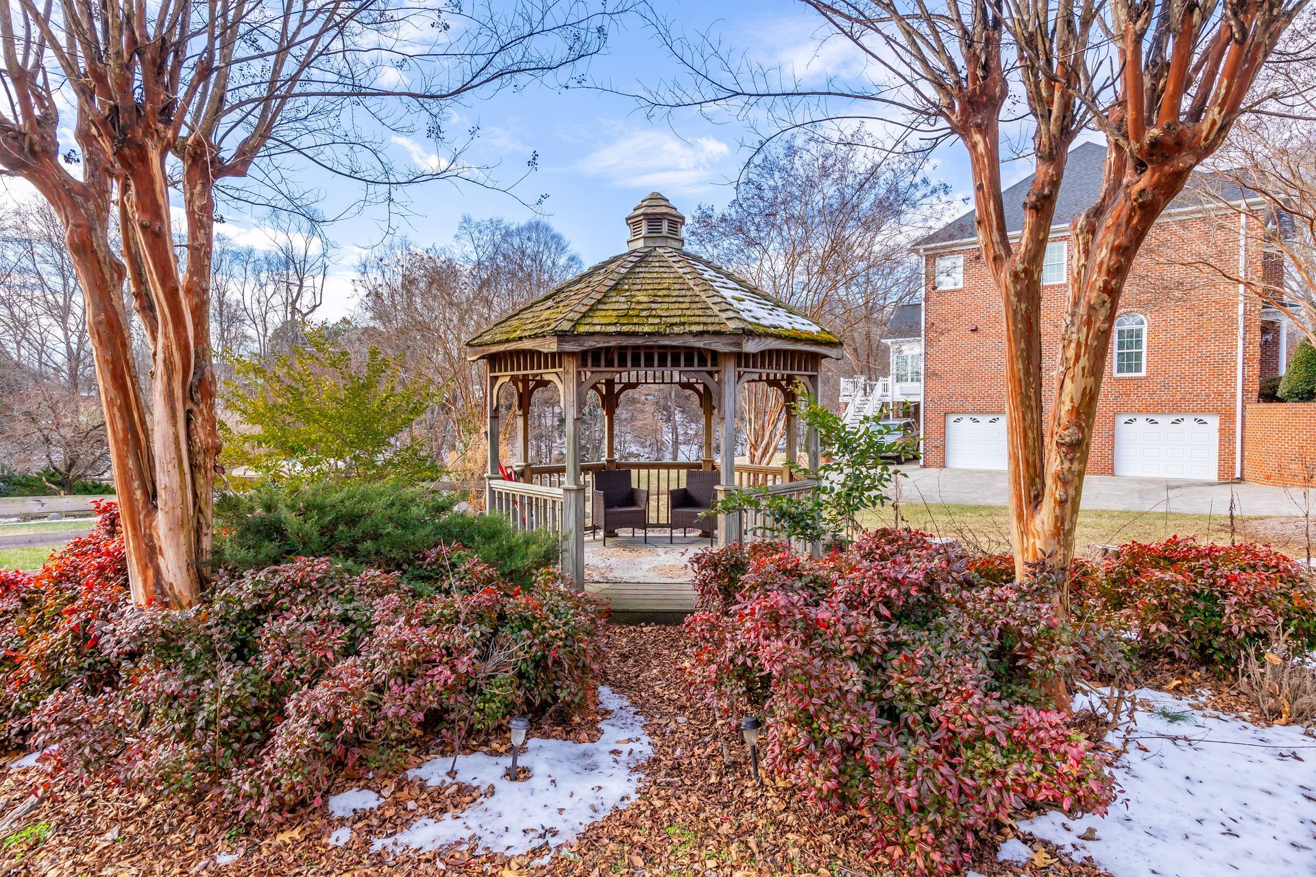 Snowy garden with a wooden gazebo, pink shrubs, and tall trees in front of a brick building.