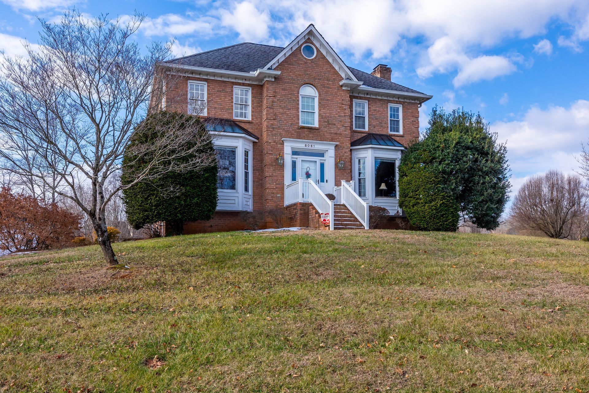 Brick two-story house with white trim and front steps on a grassy hill under a blue sky