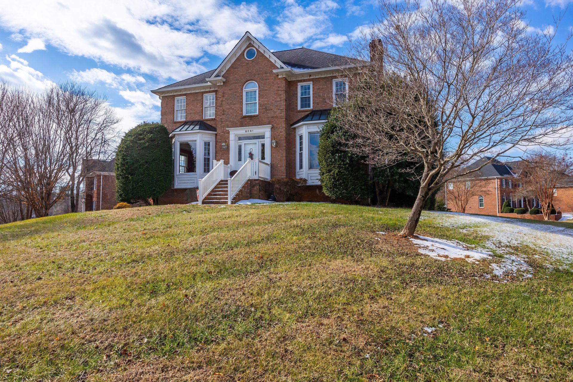 Brick colonial house on a grassy hill with white trim, bare trees, and a partly cloudy sky