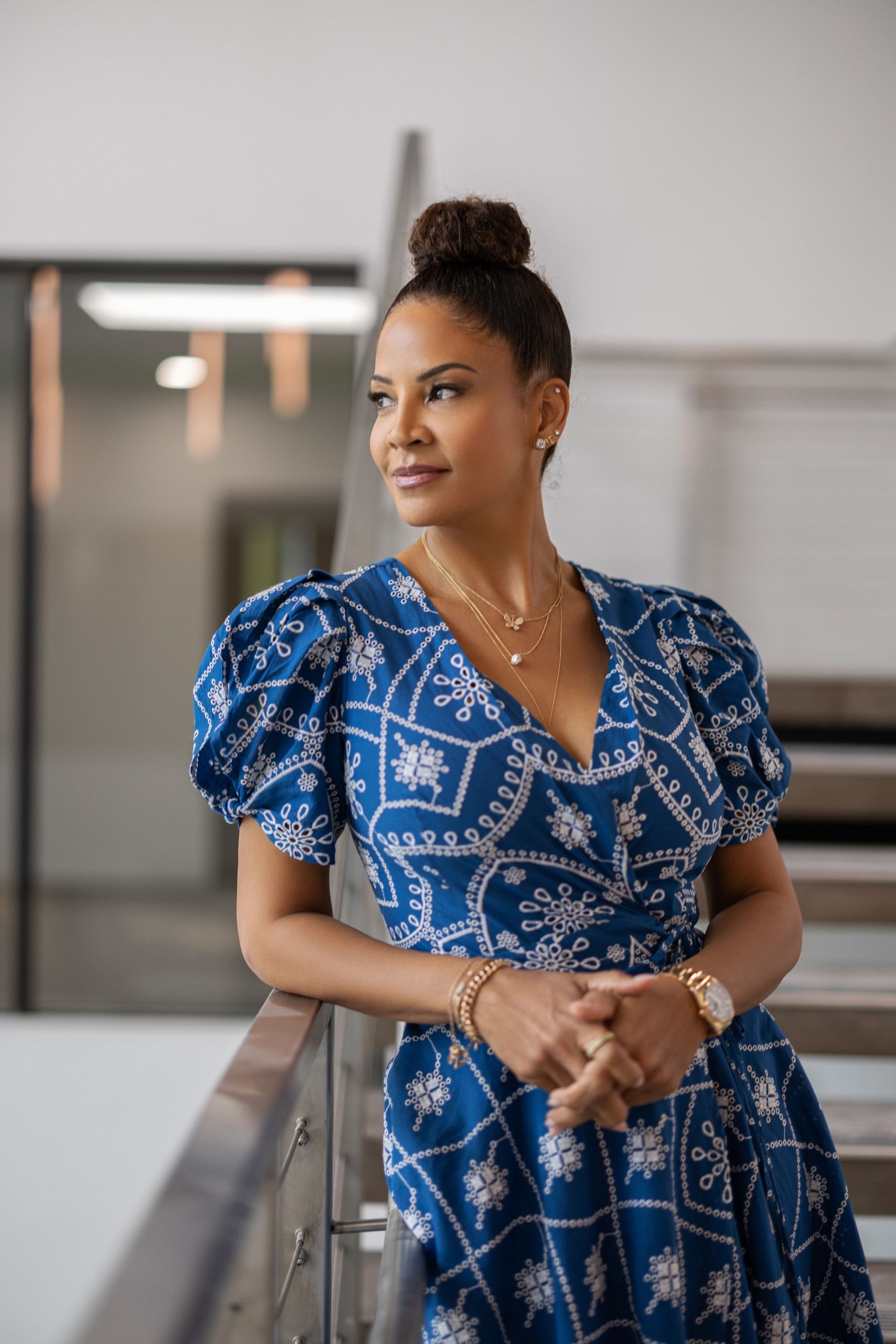 Woman in a blue patterned dress leans on a railing, looking to her right. Indoors, natural light.