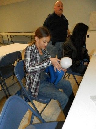 Woman inflating a balloon, supervised by a man, indoors at a table, another person in the background.