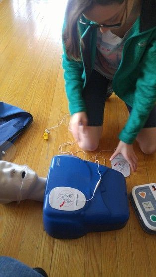 Person kneeling, training with a blue AED on a wooden floor, placing pads on a mannequin.