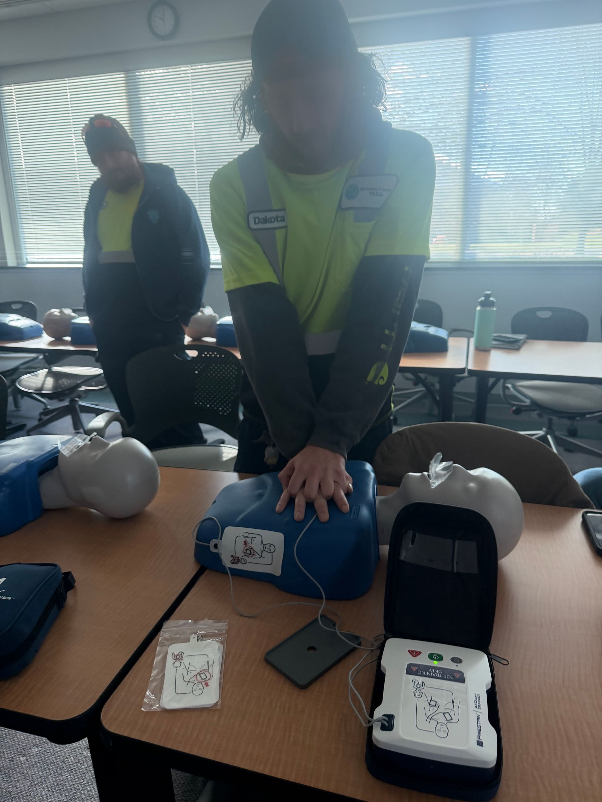Person practicing CPR on a training dummy, with an AED. Another person observes. Indoor setting.