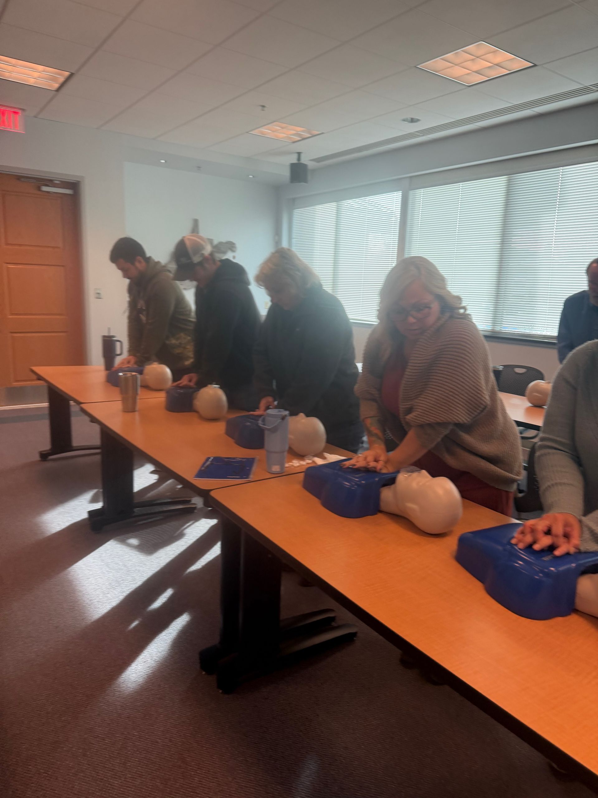 People practicing CPR on mannequins at a table in a classroom setting.
