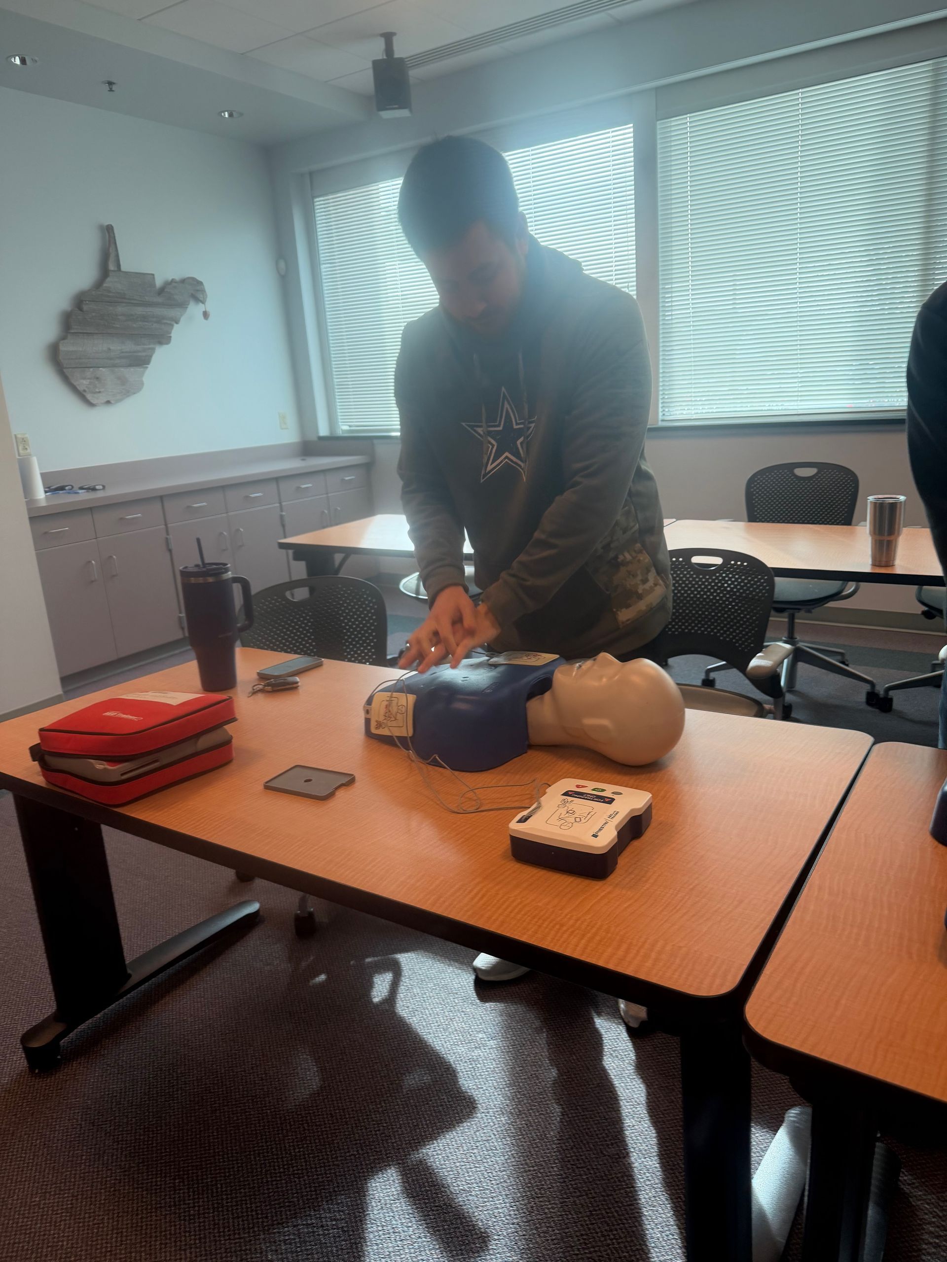 Person practicing CPR on a dummy, with an AED and emergency kit on a table in a classroom.