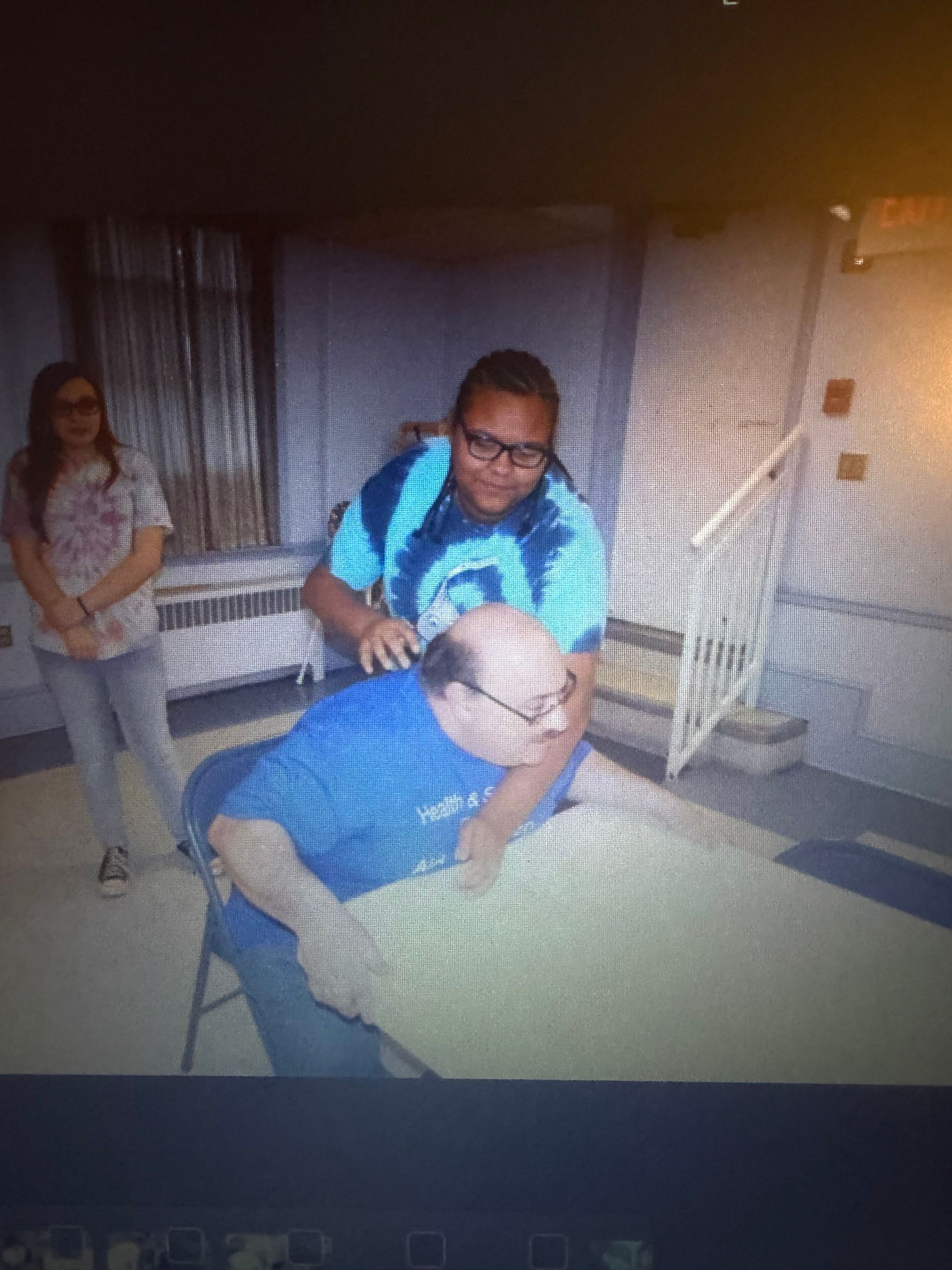 Man getting a back massage from a person in a tie-dye shirt. Another person watches from the side.