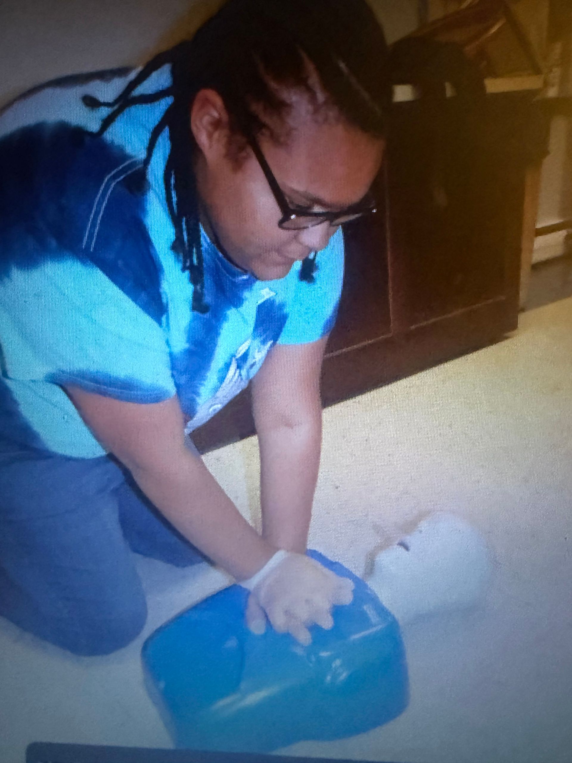 Person practicing CPR on a training dummy, pressing on its chest; blue and white tie-dye shirt.