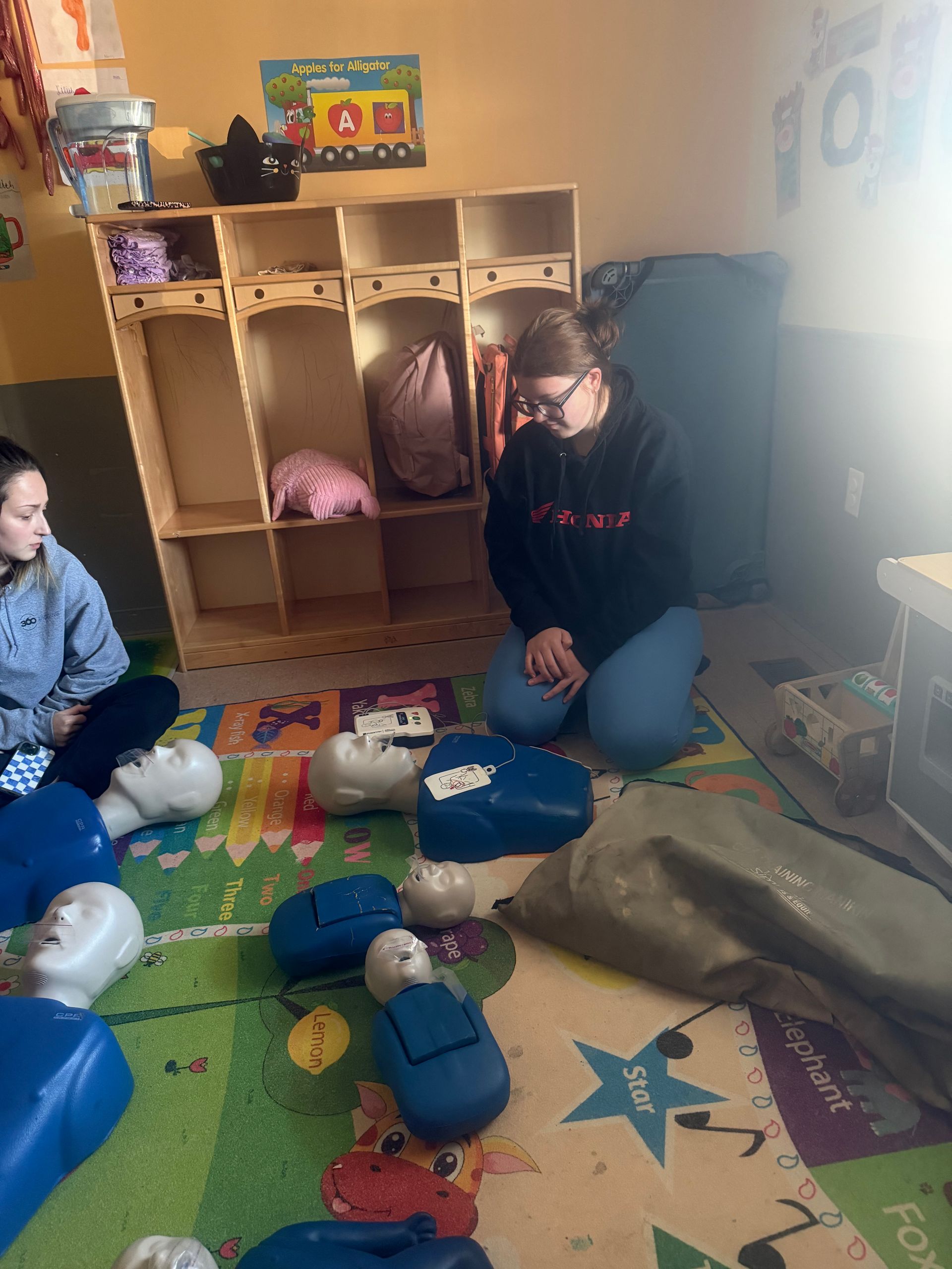 CPR training: Woman on knees near mannequins, others seated. Indoor setting, colorful rug.