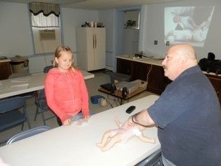 Man demonstrating infant care to a girl, using a doll in a classroom.