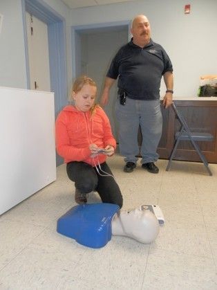 Girl kneeling, practicing with CPR dummy under supervision of a man in a room.