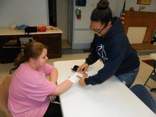 Person wrapping bandage on another's wrist at a table. Pink shirt, blue hoodie, indoor setting.