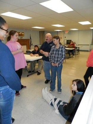People in a room, some looking at a person lying on the floor. One man gestures.