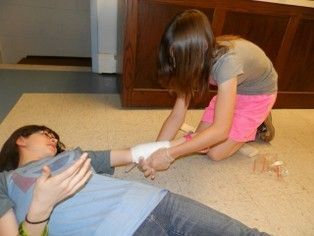 Girl bandaging another girl's arm on the floor; tan floor and wooden cabinet in background.