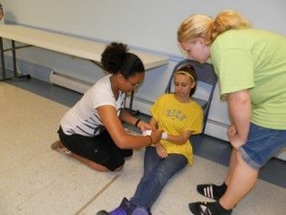 Woman kneeling, bandaging another woman's wrist; a third woman looks on indoors.