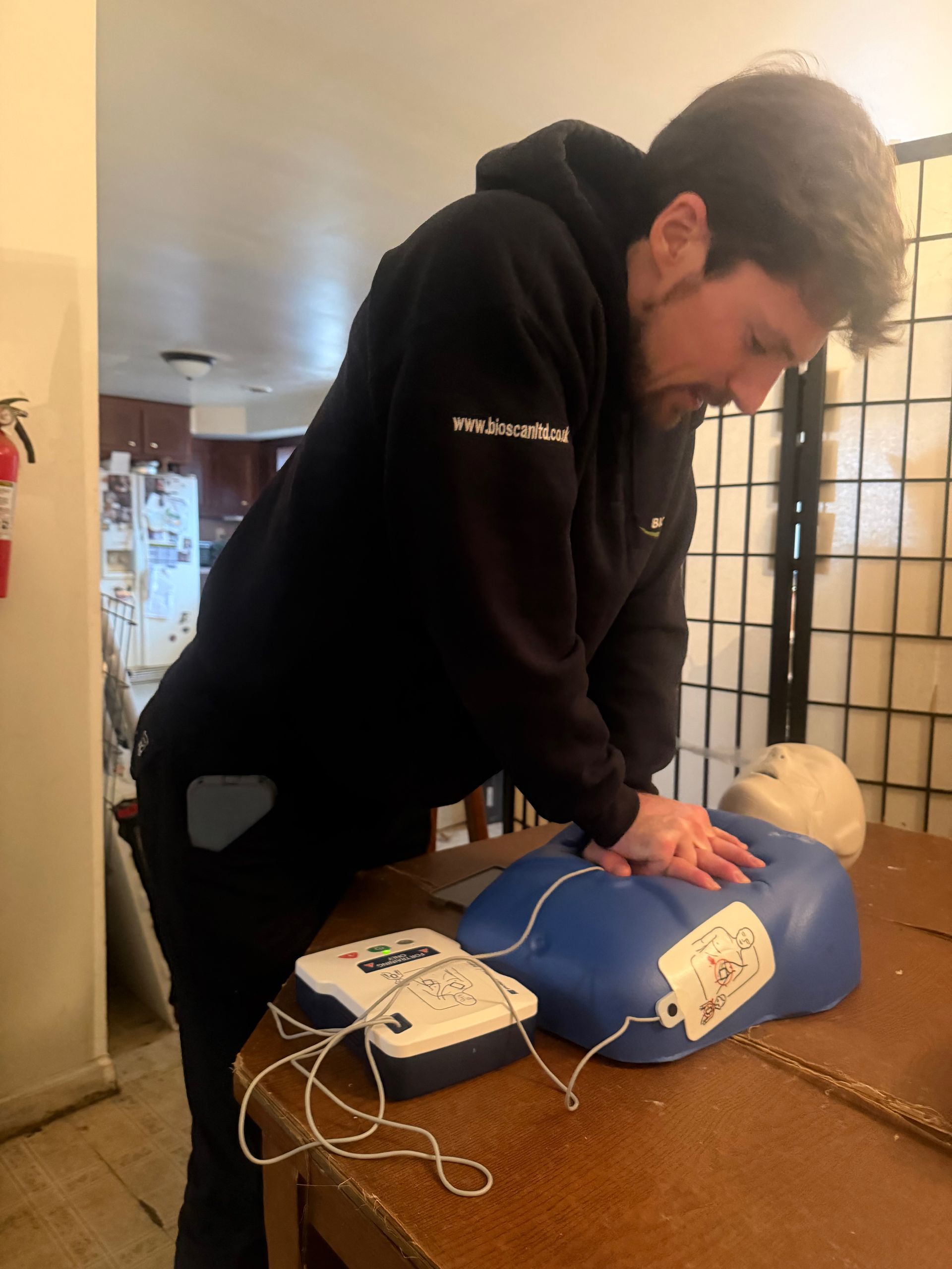 Man in black hoodie practicing CPR on a training dummy, with an AED nearby.