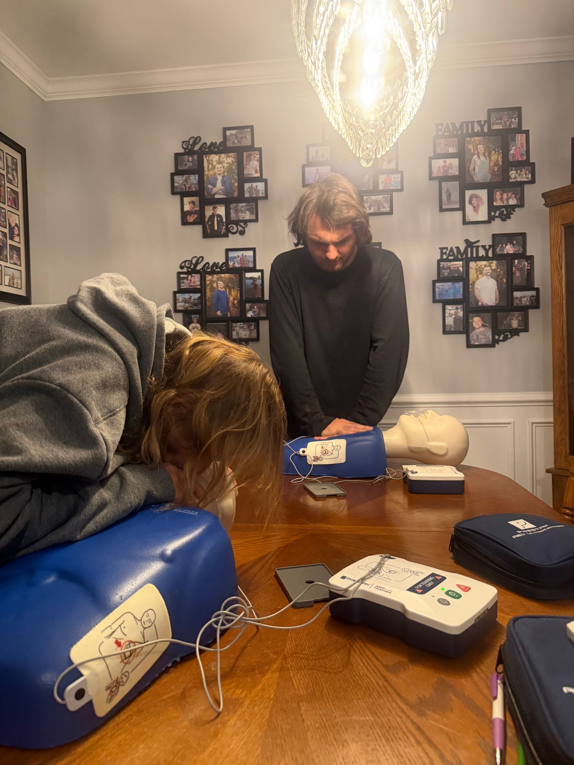 Two people practicing CPR on training dummies indoors, with an AED on the table.