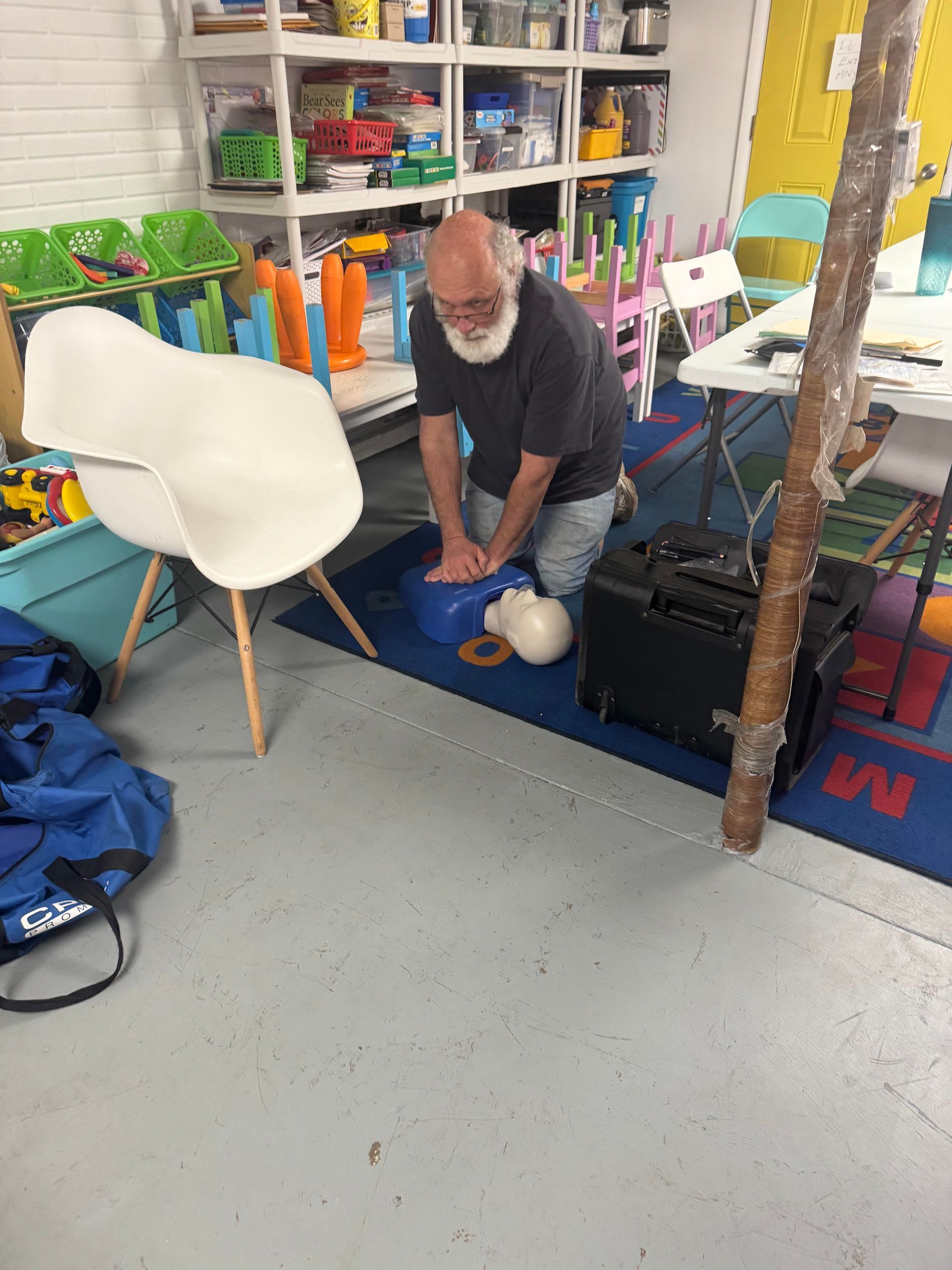 Man practicing CPR on a training mannequin in a room with shelves and a white chair.