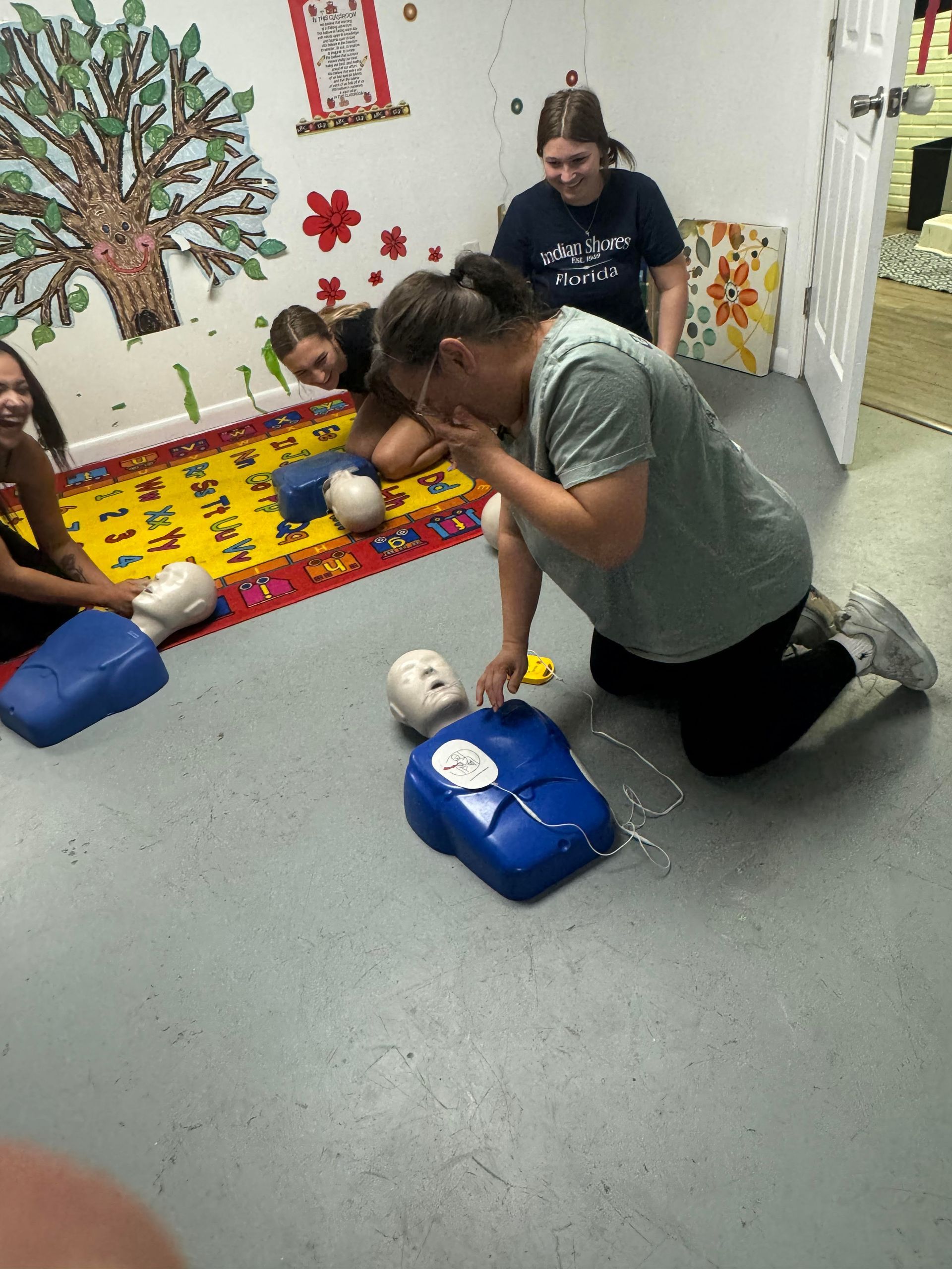 People practicing CPR on mannequins in a classroom. One woman kneels, hands on mannequin. Others observe.